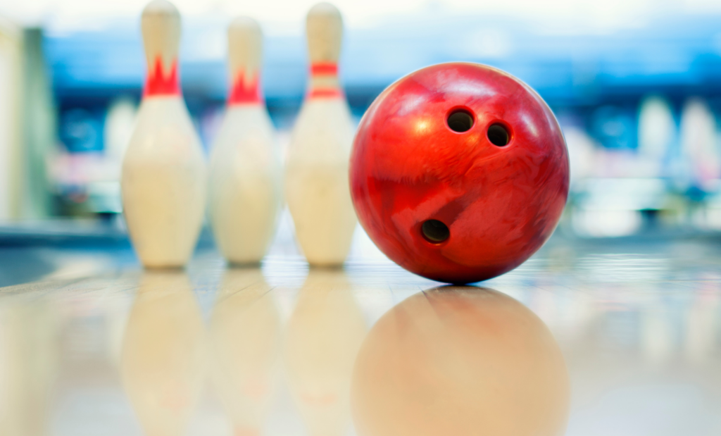 A red bowling ball and three pins sit on a brightly-lit bowling lane.