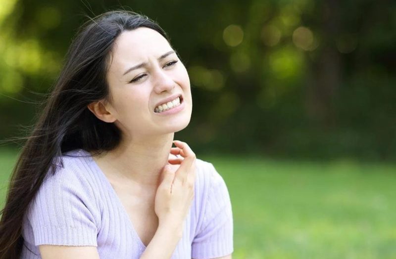 Young, dark-haired woman scratches neck while sitting outside.