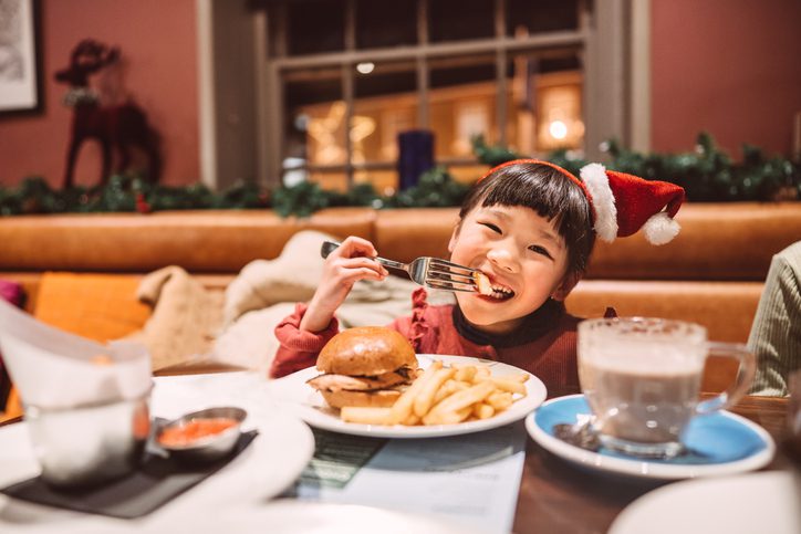 Lovely little girl in Santa hat headband smiling joyfully at the camera while enjoying burger chips in a restaurant.