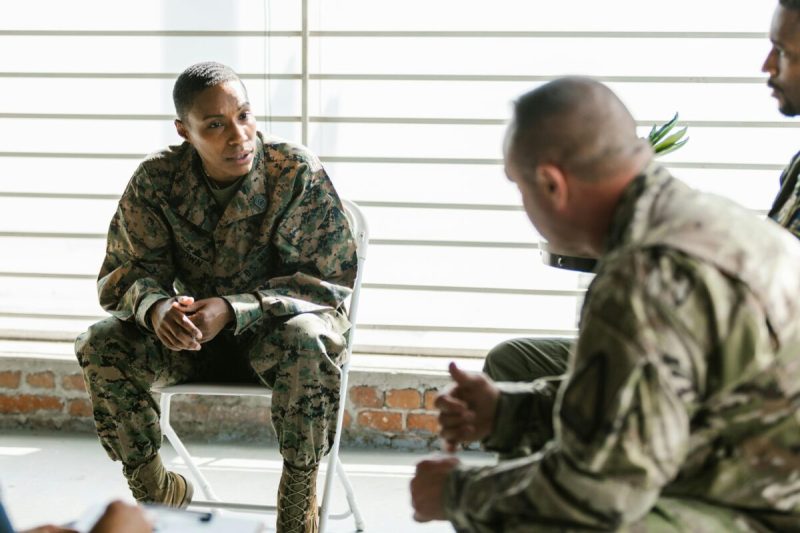 US Army woman sits on a chair while talking to several other of her comrades.