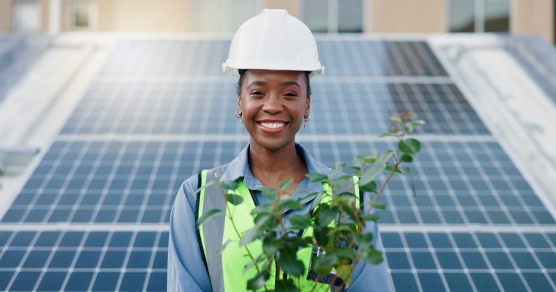 Photo of a Black woman in a construction uniform smiling and holding a plant while standing in front of a grid of solar panels.
