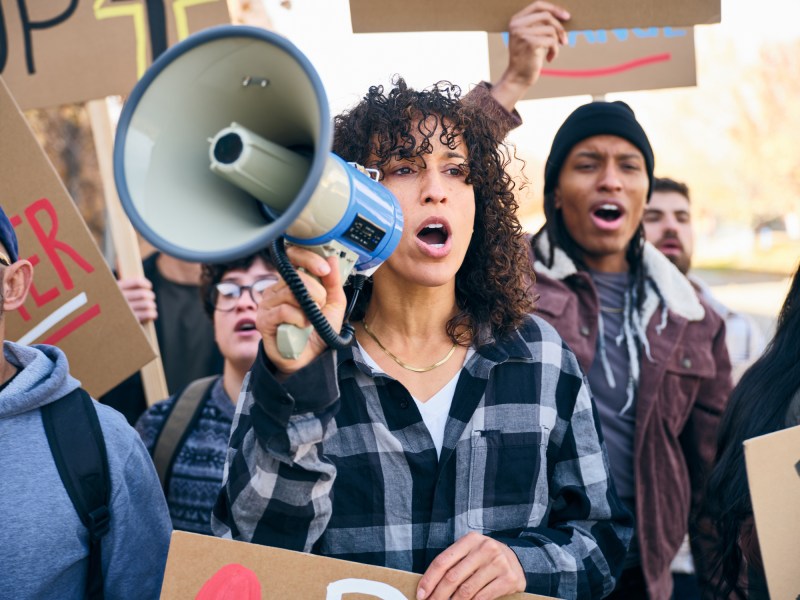 A diverse group of adults gathered outdoors holding handmade signs while a woman speaks through a megaphone during a positive community rally.